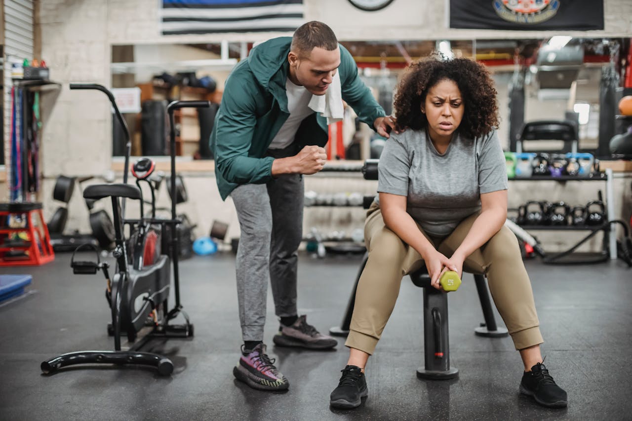 Full body of male fitness instructor in sportswear supporting tired black woman sitting with dumbbell on bench in gym